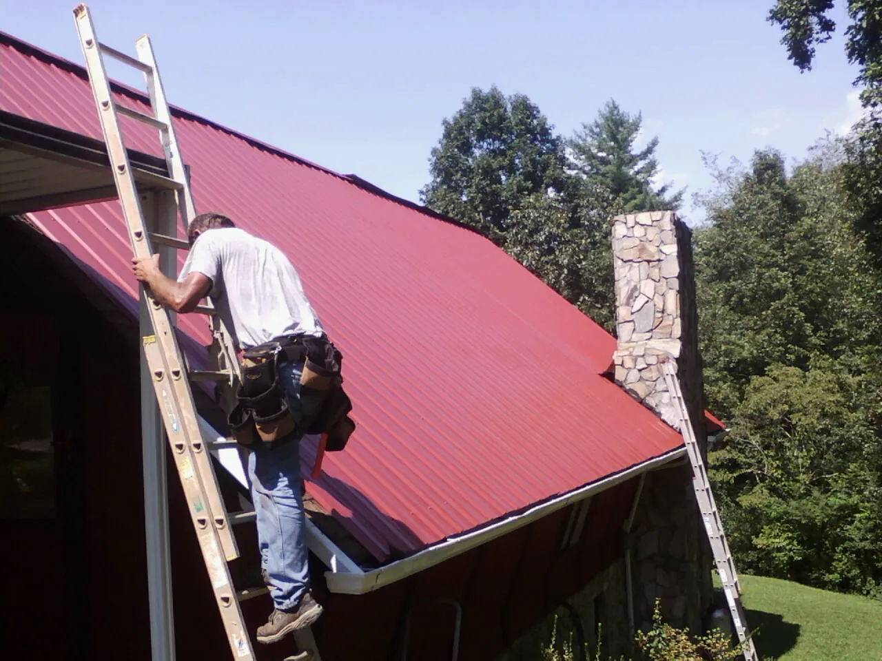 Roofer on a ladder beside a red metal roof and stone chimney — Soffit & Fascia Repair by Roofing Brothers in Turbotville